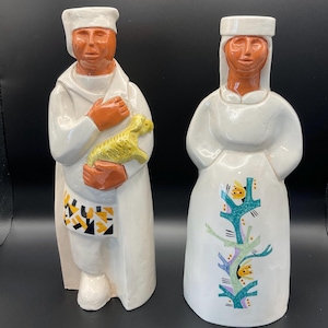 May include: Two ceramic figurines. One is a figure of a person in a white robe holding a yellow lamb. The other is a figure of a person in a white dress with a tree design. Both have brown skin and white head coverings.