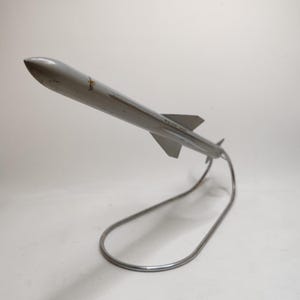 May include: A silver-colored model rocket with fins and a pointed nose, mounted on a curved metal stand. The rocket has a sleek, aerodynamic design, suggesting a focus on speed and flight. The stand provides a stable base for display.