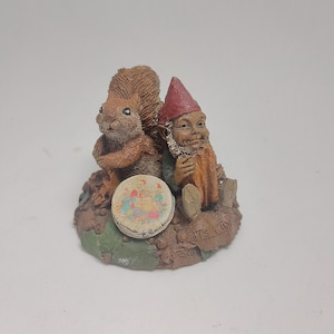 May include: A detailed figurine of a brown squirrel and a gnome with a red hat. The gnome is seated, and the squirrel stands beside him. A small, round tin with a colorful design sits in front of them. The base is textured with leaves and the words "IT'S A TIN".
