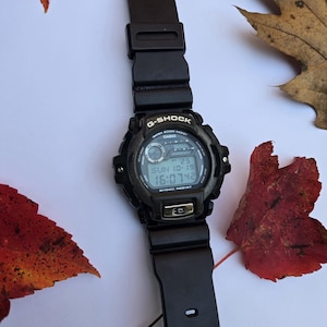 May include: Black G-Shock digital watch with a black band. The watch face displays the date and time. The watch is surrounded by red and brown autumn leaves.