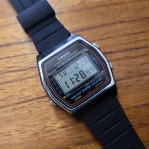 Puede incluir: Un reloj digital Casio con una correa de goma negra. La esfera del reloj muestra la hora y la fecha. El reloj tiene un bisel plateado y botones negros. El texto "CASIO" es visible en la esfera.