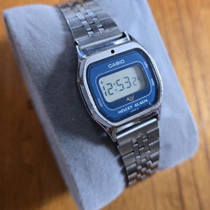 Könnte beinhalten: Eine silberfarbene Casio Digitaluhr mit blauem Zifferblatt, das die Uhrzeit 12:53 anzeigt. Die Uhr hat ein rechteckiges Zifferblatt mit dem Markennamen "CASIO" oben. Das Armband besteht aus silberfarbenen Metallgliedern.