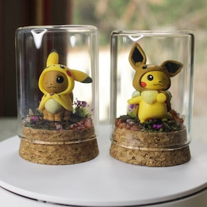 Op de afbeelding: Twee glazen terrariums met elk een gele Pikachu-figuur in een kostuum. De figuren staan op een kurken voet met planten en mos. De terrariums staan op een wit oppervlak.