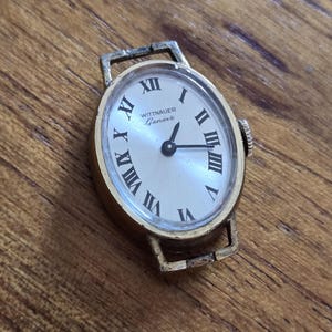 May include: A vintage gold-toned wristwatch with an oval face. The watch face is a light color with black Roman numerals and hands. The brand name "WITTNAUER Geneve" is printed on the watch face. The watch has a rectangular band attachment.