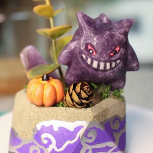 Puede incluir: Una maceta decorativa con una figura de Gengar morado, una calabaza naranja y una piña. La maceta tiene una parte superior color arena y una base con diseños morados y plateados. La vegetación y una planta suculenta completan el diseño.