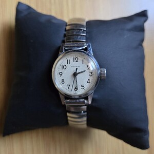May include: A vintage silver-toned wristwatch with a white face and black numerals, marked "SIGNAL." The watch has a stretch band and a small crown. The watch is displayed on a black pillow.
