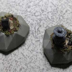 Puede incluir: Dos jardineras grises de forma geométrica. Una presenta una casa en miniatura con un tejado oscuro y la otra una tortuga en miniatura y una figura con sombrero. Ambas jardineras tienen musgo y pequeñas piedras.