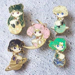 May include: Five gold-colored enamel pins featuring anime characters from the Sailor Moon franchise. Each pin depicts a different character with a unique pose and background. The characters are all wearing colorful outfits and have sparkling eyes. The pins are perfect for collectors or fans of the show.