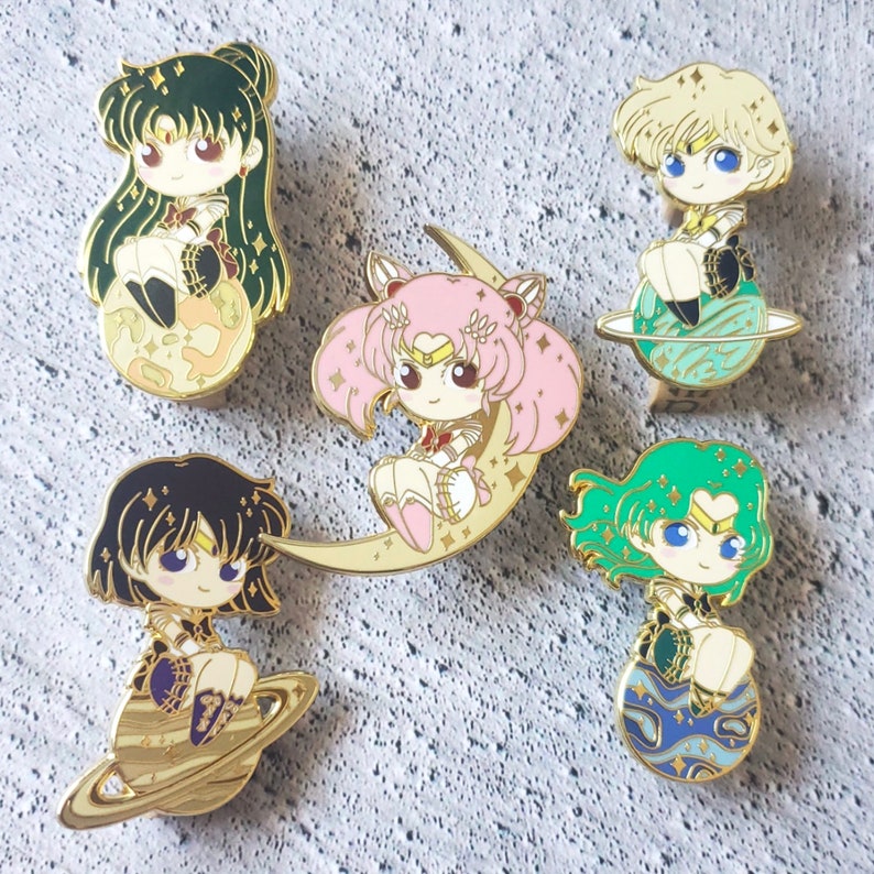 May include: Five gold-colored enamel pins featuring anime characters from the Sailor Moon franchise. Each pin depicts a different character with a unique pose and background. The characters are all wearing colorful outfits and have sparkling eyes. The pins are perfect for collectors or fans of the show.