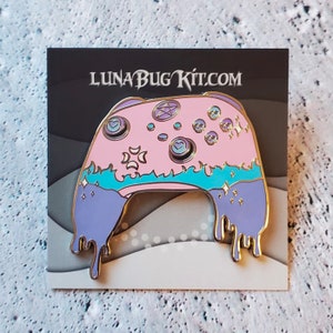 Op de afbeelding: Een roze en paarse emaille pin in de vorm van een videogamecontroller met een druppelend effect. De controller heeft een pentagram, manen, harten en schedels erop. De pin zegt "LUNA BUG KIT.COM".