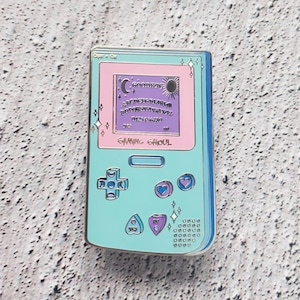 May include: A light blue and pink enamel pin shaped like a handheld video game console. The screen displays a purple Ouija board with the text "GOODBYE" and a crescent moon and sun. The text "GAMING GHOUL" is below the screen. The console has a directional pad, two heart-shaped buttons, and a Ouija planchette button.