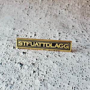May include: Gold-colored metal pin with black enamel. The pin has the text "STFUATTDLAGG" in all caps.