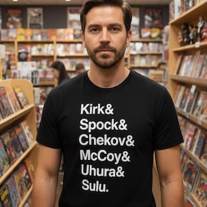 Può includere: T-shirt nera girocollo con testo bianco che elenca i nomi Kirk, Spock, Chekov, McCoy, Uhura e Sulu. La persona si trova in una libreria con scaffali di libri sullo sfondo.