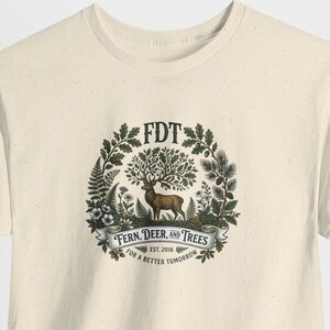 Può includere: T-shirt color crema con un'illustrazione dettagliata. Il design presenta un cervo con corna simili ad alberi, circondato da felci e fogliame. Il testo include "FDT", "Fern, Deer, and Trees" e "For a Better Tomorrow".