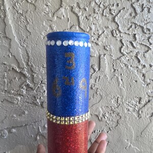 May include: A cylindrical tumbler featuring a blue glitter top section and a red glitter bottom section. The blue portion displays the numbers 3, 6, and 9 in a dark font. A row of clear rhinestones circles the rim, and a gold rhinestone band separates the colors.
