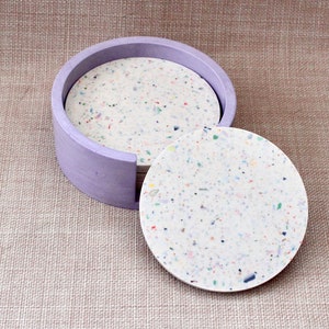 May include: A set of six round coasters with a speckled white and multi-colored design. The coasters are in a purple holder.