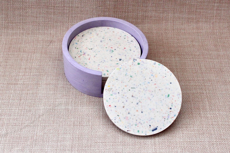 May include: A set of six round coasters with a speckled white and multi-colored design. The coasters are in a purple holder.