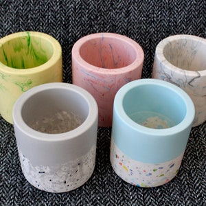 May include: Five cylindrical concrete planters in different colours and patterns. The planters are arranged in a circle on a grey and white patterned surface. The planters are made of concrete and have a smooth, polished finish. The colours of the planters are yellow, pink, grey, blue, and white.