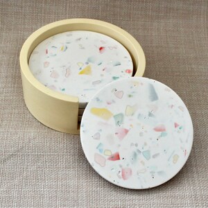 May include: Set of six round coasters with a white base and colorful flecks. The coasters are in a cream-colored holder.