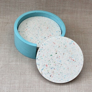 May include: Set of four round coasters in a light blue holder. The coasters are made of white speckled material with flecks of blue, red, and yellow.