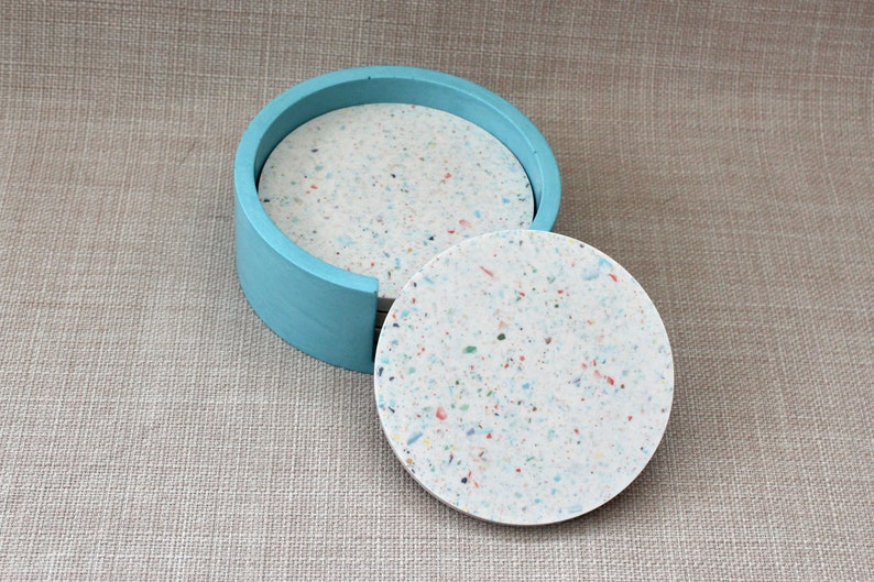 May include: Set of four round coasters in a light blue holder. The coasters are made of white speckled material with flecks of blue, red, and yellow.