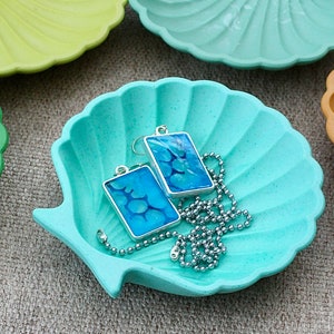 May include: Two blue resin pendant earrings with a silver chain on a light blue seashell-shaped dish.
