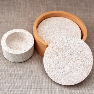 May include: Set of four round coasters with a speckled white and brown design. The coasters are stacked and have a wooden frame.