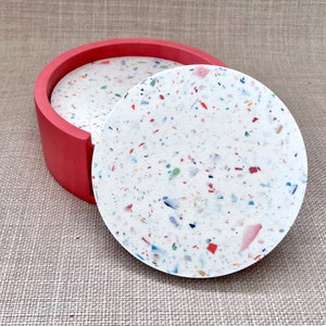 May include: Set of four round coasters with a white base and colorful flecks. The coasters are in a red holder.