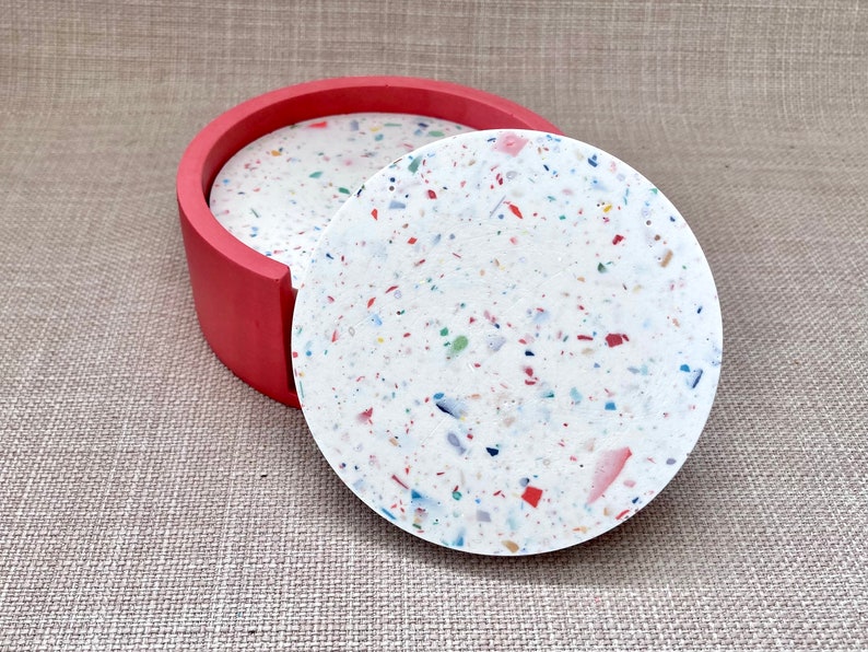 May include: Set of four round coasters with a white base and colorful flecks. The coasters are in a red holder.