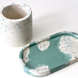 May include: A set of two concrete trays with a speckled pattern in white and blue. The larger tray is oval shaped and has a light green glaze. The smaller tray is cylindrical.