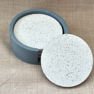 May include: Set of six round gray and white speckled concrete coasters in a gray holder. The coasters are stacked and one is pulled out.