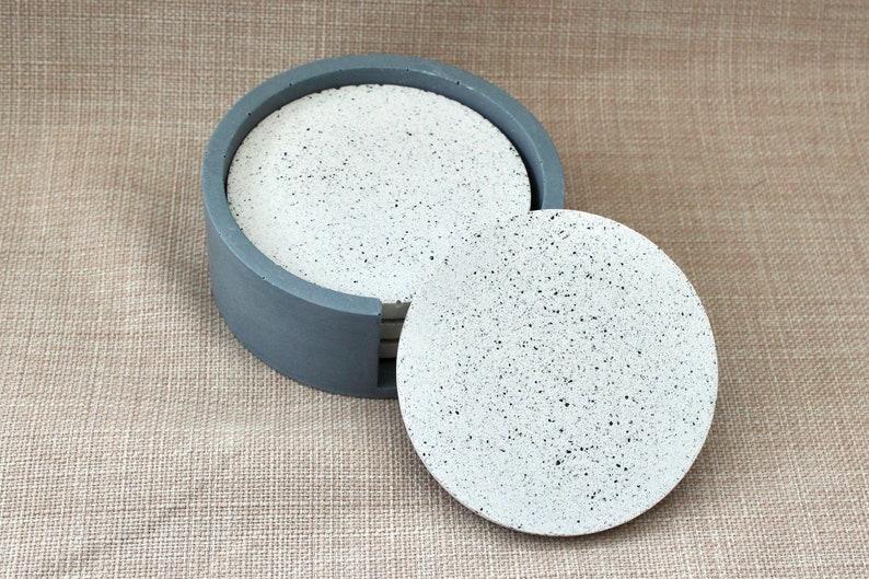 May include: Set of six round gray and white speckled concrete coasters in a gray holder. The coasters are stacked and one is pulled out.