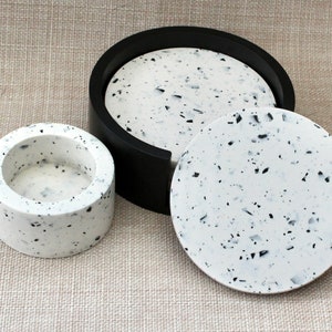 May include: Three round coasters made of white speckled resin with black flecks. The largest coaster has a black rim.