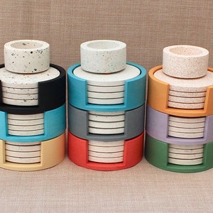 May include: Four sets of coasters in different colors. Each set has a stack of white coasters with black speckles and a matching top coaster. The coasters are stacked in a holder with a different color for each set: black, blue, red, and green.