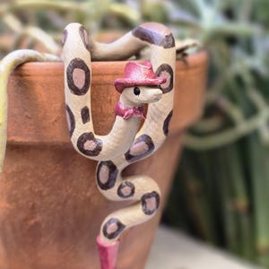 May include: A whimsical, hand-painted ceramic snake figurine. The snake is cream-colored with brown spots and a brown head. It wears a pink cowboy hat, a pink bow tie, and a pink boot. The snake is posed around a terracotta pot.