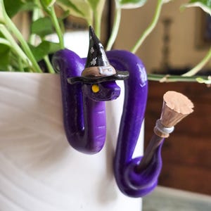 May include: A purple snake figurine wearing a black witch hat with white star accents and a brown band. The snake has yellow eyes and is holding a small brown broomstick with a tan bristle end. The figurine is curved to hook onto a white surface.