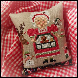 May include: A cross-stitch pillow featuring a red-dressed woman with a gingerbread cookie in her hand, surrounded by a reindeer, a snowman, a cat, and a house. The pillow has a black and white trim.