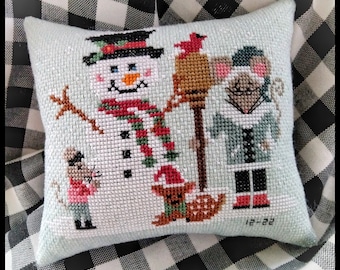 May include: A light blue cushion with a cross-stitch design featuring a snowman, a mouse, a bird, and a small dog. The design is set against a snowy background. The cushion has a date of "12-22" stitched in the bottom right corner.