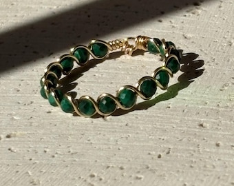 May include: A wire-wrapped ring featuring small, round, faceted dark green beads secured by a thin gold-colored metal wire. The ring is circular with a twisted wire design that holds each individual green bead in place along the entire band.