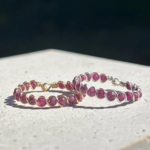 May include: Two rings with pink gemstones, each ring is wire-wrapped. One ring has gold-coloured wire, the other silver. The rings are displayed on a white surface with a dark background. The gemstones are small and round.