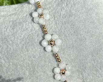 May include: A delicate bracelet featuring white flower-shaped beads and gold-colored accents. The bracelet is displayed on a gray, textured surface, with a blurred green background of grass and foliage.
