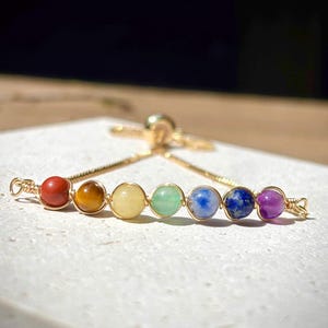 May include: A gold-tone bracelet with seven gemstone beads in chakra colours: red, orange, yellow, green, blue, indigo, and purple. The bracelet has an adjustable design and is delicate in appearance.
