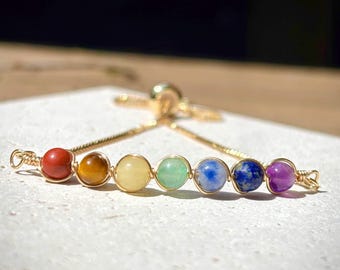 May include: A gold-tone bracelet with seven gemstone beads in chakra colours: red, orange, yellow, green, blue, indigo, and purple. The bracelet has an adjustable design and is delicate in appearance.