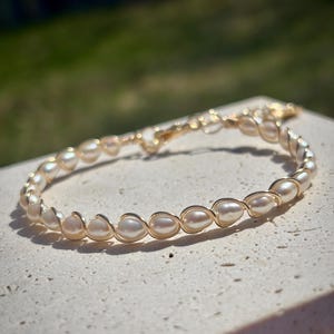 Peut inclure: Un bracelet composé d'une série de petites perles blanc cassé en forme de goutte, chacune enveloppée individuellement dans un fil métallique doré. Le fil forme un motif de boucle continu et répétitif autour de chaque perle, les reliant.
