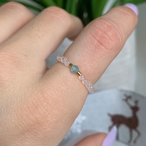 May include: A delicate ring made of small white beads with a single light blue gemstone in the centre. The ring is worn on a finger.