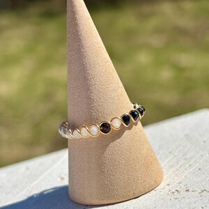 May include: A gold-toned wire ring featuring a series of small, round gemstones in alternating colors of clear, white, brown, and black. The ring is displayed on a beige, cone-shaped stand against a blurred green background.