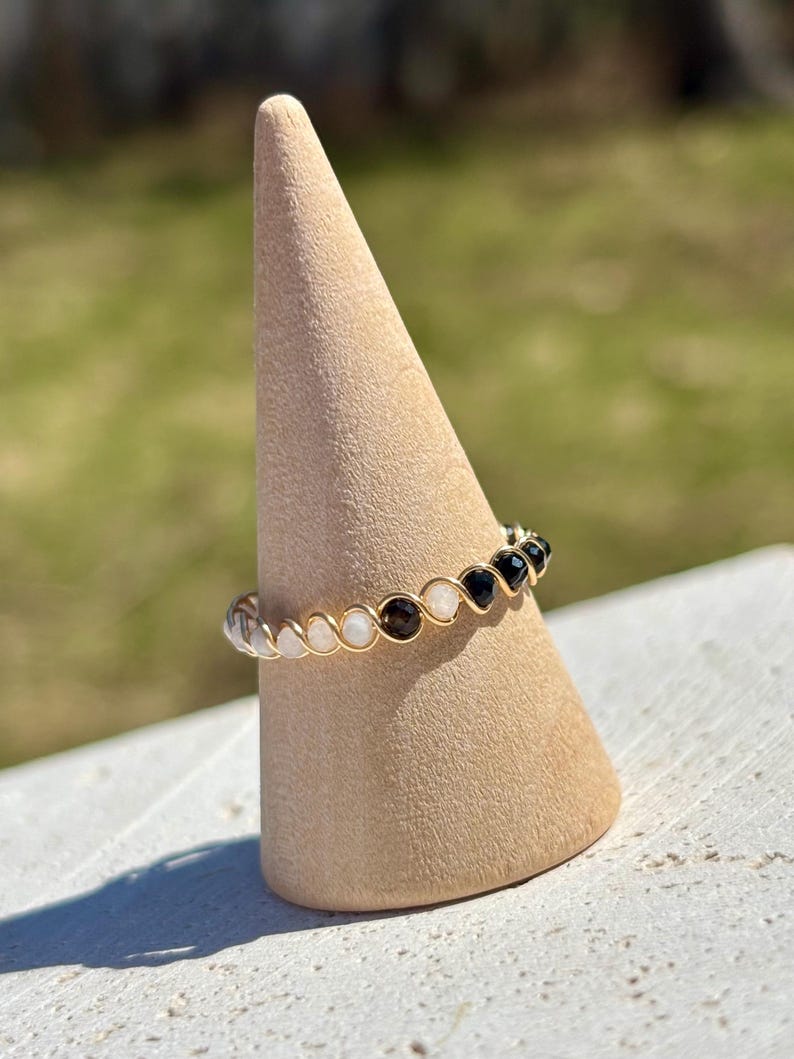 May include: A gold-toned wire ring featuring a series of small, round gemstones in alternating colors of clear, white, brown, and black. The ring is displayed on a beige, cone-shaped stand against a blurred green background.