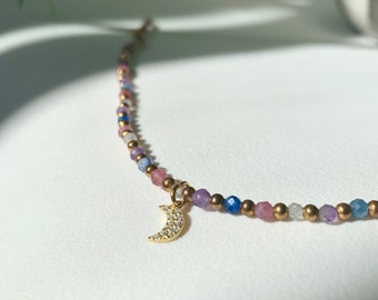 Peut inclure: Un collier de perles délicat avec un pendentif en forme de croissant de lune doré incrusté de petites pierres claires. Le collier est composé d'une alternance de petites perles dorées métalliques et de perles en pierre gemme facettées roses, bleues et violettes.