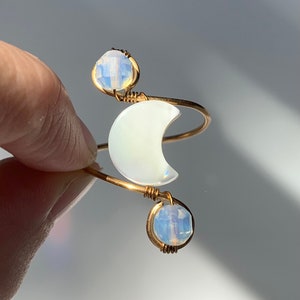 Könnte beinhalten: Ein goldfarbener, drahtgewickelter Ring mit einem weißen Halbmond-Anhänger und zwei schillernden Opal-Steinen.