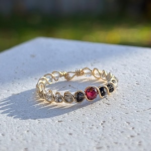 Peut inclure: Une bague filaire dorée ornée de pierres transparentes et gris foncé, ainsi qu'une pierre rouge centrale. La bague est posée sur une surface blanche avec un arrière-plan vert flou, mettant en valeur un design délicat et artisanal.